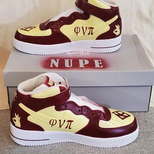 May include: High-top sneakers in cream, burgundy, and yellow with white soles. The sneakers feature the Greek letters Φ, Ν, and Π, along with a hand gesture and the word "NUPE" on the box. A white t-shirt with matching Greek letters is visible.