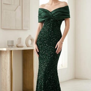 Op de afbeelding: Een vloerlange, donker smaragdgroene japon met een nauwsluitend silhouet bedekt met glinsterende pailletten. Het lijfje heeft een off-shoulder halslijn met een gladde, gedrapeerde satijnen overlay die de bovenarmen en borst bedekt.