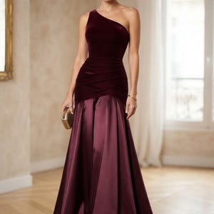 May include: A floor-length gown in deep burgundy featuring a velvet bodice with a single shoulder strap and a ruched waistline. The skirt is crafted from a smooth, satin-like fabric that flares out from the hips to the floor in a full silhouette.