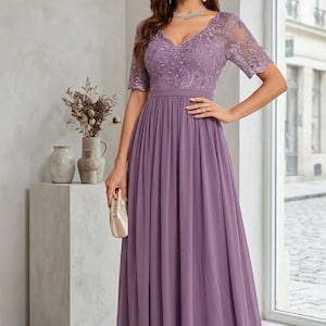 May include: A long, lavender-colored formal dress with a V-neckline and short sleeves. The bodice features intricate lace detailing, and the skirt flows to the floor. A small, cream-colored clutch is held in the hand.