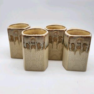 May include: A set of four hexagonal ceramic mugs with a cream-colored glaze and brown drip accents. Each mug features a raised, embossed geometric pattern on the exterior surface, with a smooth interior and a distinct, angular rim shape.