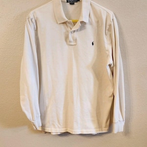 May include: A long-sleeve cream-colored polo shirt featuring a collar, a two-button placket, and a dark blue embroidered logo on the left chest. The interior neck label displays the text Polo by Ralph Lauren with the size L clearly visible.