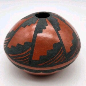 May include: A small, round, terracotta-coloured pottery vessel with a smooth, polished finish. The surface is decorated with matte black geometric patterns, including stepped triangular shapes and curved lines encircling the base.