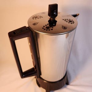 Puede incluir: Una cafetera percoladora de metal plateado con asa, base y pomo de tapa de plástico marrón oscuro. La tapa circular presenta un patrón floral negro compuesto por pequeños puntos dispuestos en forma de estrella.
