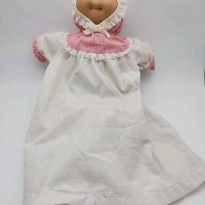 May include: A soft-sculpture doll with a tan fabric face, blue eyes, and yarn hair. The doll wears a white nightgown with pink gingham accents, a matching bonnet with white lace trim, and a small white ribbon bow tied at the neck.