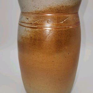 May include: A cylindrical stoneware vase featuring a two-tone glaze with a light gray upper section and a warm, speckled brown lower body. A horizontal band with subtle, incised decorative markings encircles the upper portion of the vase.