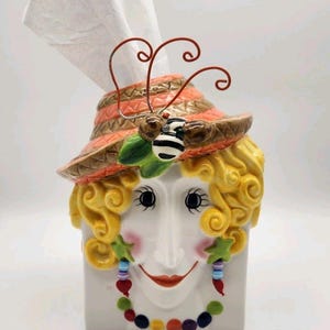 May include: A ceramic tissue box cover shaped like a face with curly yellow hair, wearing an orange and brown striped hat with a bee accent. The face features pink cheeks, star-shaped earrings with dangling beads, and a colorful beaded necklace.