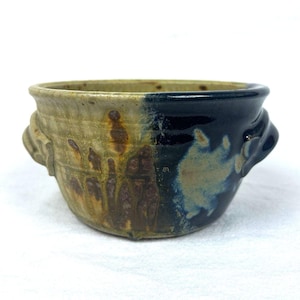 May include: A ceramic bowl with two side handles, featuring a two-tone glaze. One side is a textured tan with brown drip patterns, while the other side is a smooth, deep navy blue. The bowl has a wide, open top and a rounded base design.