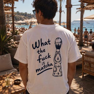 May include: A white short-sleeved t-shirt featuring a dark blue graphic on the back. The design includes the text "What the fuck is matcha" written in a casual script font next to a detailed illustration of a decorative tequila bottle.