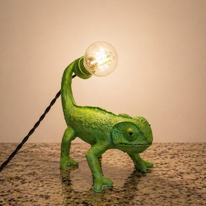 Può includere: Una stravagante lampada a forma di camaleonte verde con una lampadina trasparente. La lampada ha una superficie strutturata e un cavo nero. La lampadina è tenuta dal collo del camaleonte, creando un'illuminazione unica.