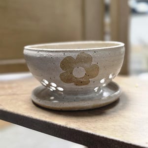May include: A speckled off-white ceramic bowl with a wide, rounded rim and a series of small circular cutouts around the base. The exterior features a single, large, tan-colored flower design with a central circle, resting on a matching saucer.