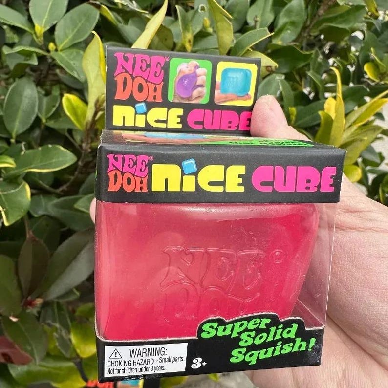 May include: A red Nee Doh Nice Cube toy in a clear plastic container. The box is black with the words "Nee Doh Nice Cube" in pink, yellow, and blue. The bottom of the box reads "Super Solid Squish!" and has a warning label.