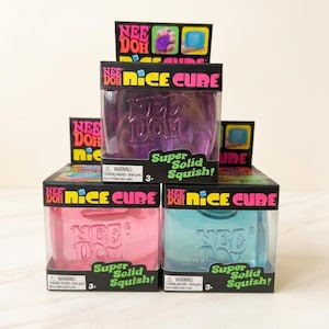 May include: A stack of Nee Doh Nice Cube toys in purple, pink, and blue. Each box has the Nee Doh logo and the words "Super Solid Squish!" The boxes are black with colourful accents and a clear plastic window.