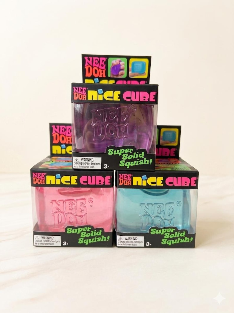 May include: A stack of Nee Doh Nice Cube toys in purple, pink, and blue. Each box has the Nee Doh logo and the words "Super Solid Squish!" The boxes are black with colourful accents and a clear plastic window.