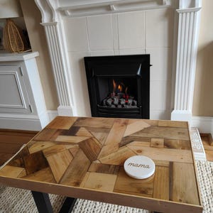 May include: A rectangular coffee table with a mosaic wood top and black metal legs. A white coaster with the word "mama" is on the table. The table sits on a cream-colored rug in front of a fireplace.