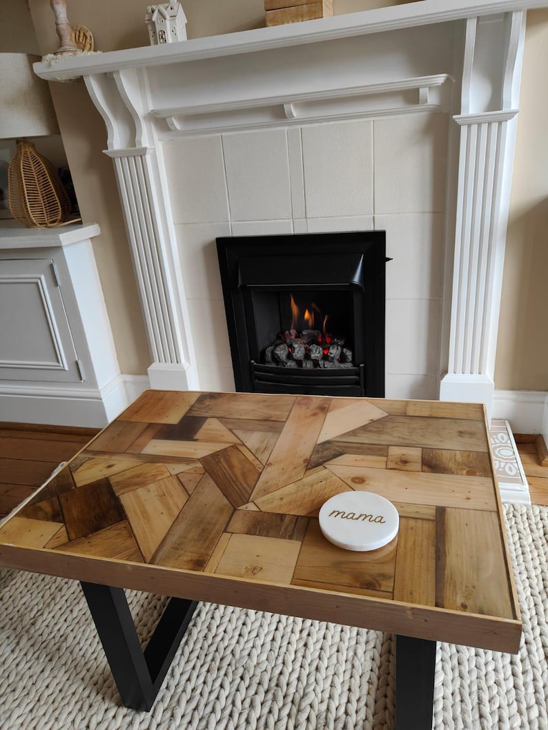 May include: A rectangular coffee table with a mosaic wood top and black metal legs. A white coaster with the word "mama" is on the table. The table sits on a cream-colored rug in front of a fireplace.
