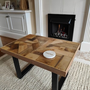 May include: A rectangular coffee table with a wooden mosaic top and black metal legs. The table has a light brown wooden frame and a white coaster with the word "mama" on it. The table is 48 inches long and 24 inches wide.