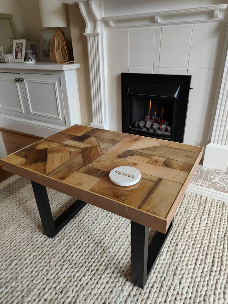 May include: A rectangular coffee table with a wooden mosaic top and black metal legs. The table has a light brown wooden frame and a white coaster with the word "mama" on it. The table is 48 inches long and 24 inches wide.