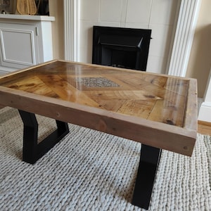 May include: Rectangular coffee table with a wooden top featuring a chevron pattern and a clear, glossy finish. The table has a light brown wooden frame and black metal legs. The table is approximately 122 cm long and 61 cm wide.