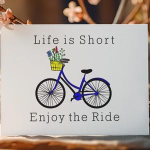 May include: A white card with the text "Life is Short Enjoy the Ride". The card features a blue bicycle illustration with a yellow basket filled with flowers. The bicycle has black wheels and a black seat.