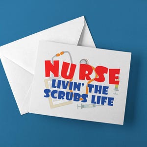 May include: A white greeting card with a matching envelope featuring the red text NURSE and blue text LIVIN' THE SCRUBS LIFE. The design includes illustrations of a stethoscope, a clipboard, and two syringes arranged behind the bold lettering.