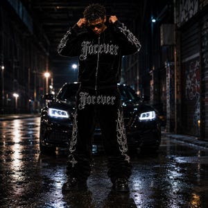 Puede incluir: Chándal de terciopelo negro con la palabra "Forever" en fuente gótica en la chaqueta y el pantalón. La chaqueta tiene cremallera y el pantalón un diseño de llama. El conjunto está adornado con pedrería plateada. La persona lleva gafas de sol negras.