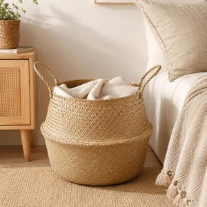 Può includere: Un cesto rotondo in erba marina intrecciata di colore beige naturale, dotato di due robuste maniglie intrecciate sui lati. Il cesto presenta una base ampia e arrotondata che si restringe verso l'alto e contiene del morbido tessuto bianco.