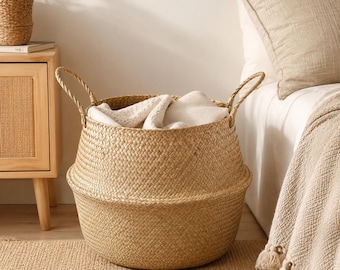 May include: A round, woven seagrass basket with a natural tan colour, featuring two sturdy braided handles on the sides. The basket has a wide, rounded base that tapers slightly towards the top and contains a soft, folded white fabric inside.