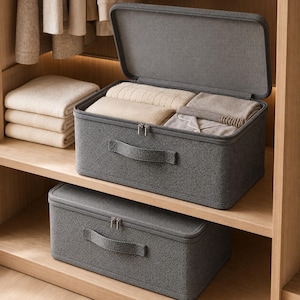 May include: Two rectangular dark grey fabric storage bins with lids and handles. The top bin is open, revealing folded beige and grey clothing items inside. The bins feature a textured weave finish and dual zipper closures for secure storage.