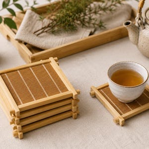 May include: A stack of square bamboo coasters with a matching coaster holding a white teacup filled with tea. A wooden tray with a linen napkin and sprigs of greenery sits in the background, along with a teapot.