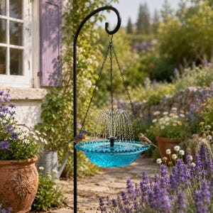 Puede incluir: Fuente colgante para pájaros con un cuenco de cristal azul y un soporte de metal negro. La fuente rocía agua hacia arriba. Dos pequeños pájaros se posan en el borde del cuenco. El fondo incluye un jardín con flores.