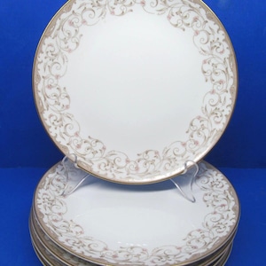 May include: A stack of five round white porcelain dinner plates featuring a delicate gold rim and a decorative border pattern of light brown swirling filigree vines accented with tiny pink floral buds along the outer edge of each plate.