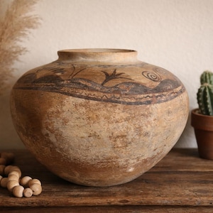 May include: A large, round, antique-style earthenware vase with a wide opening. The vase has a light brown base color with a decorative band of dark brown and black painted designs near the top. A small cactus in a terracotta pot sits nearby.