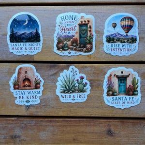 May include: Six decorative stickers featuring Southwestern themes. Designs include a night landscape with text 'SANTA FE NIGHTS MAGIC & QUIET', a doorway with 'HOME IS WHERE THE HEART IS', a hot air balloon with 'RISE WITH INTENTION', a fireplace with 'STAY WARM BE KIND', desert plants with 'WILD & FREE', and a blue door with 'SANTA FE STATE OF MIND'.