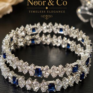 May include: Two silver-toned bangles featuring a repeating pattern of clear marquise-cut stones arranged in floral clusters, interspersed with rectangular deep blue stones. The Noor & Co logo and the text Timeless Elegance appear above the jewelry.