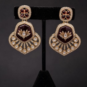 May include: A pair of gold-toned earrings with a dark red center and a floral design. The earrings have a square top with red accents and a teardrop-shaped bottom with clear stone embellishments. The earrings are displayed on a black stand.