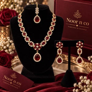 May include: A gold-toned jewelry set featuring a necklace, matching drop earrings, and a maang tikka. Each piece is adorned with deep red, pear-shaped and round stones surrounded by small clear crystals. The set is branded with the text Noor n co.