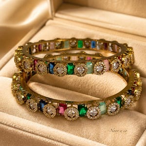 May include: Two gold-tone metal bangles featuring an alternating pattern of rectangular multi-colored gemstones and circular clear stones. The jewelry pieces are set with emerald green, royal blue, pink, and light blue stones, with the text Noor n co.