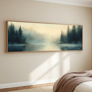 Puede incluir: Una pintura de paisaje rectangular enmarcada representa una serena escena lacustre. Árboles verde oscuro bordean la orilla del agua, con una atmósfera brumosa y una suave luz dorada en el fondo. El marco es de color madera clara.