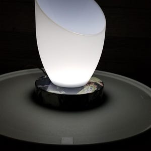 May include: A table lamp featuring a translucent white, tulip-shaped glass shade that emits a soft glow. The shade is mounted on a circular base with a dark, multi-colored marbled pattern that includes veins of black, white, and earthy tones.