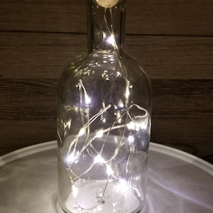 May include: A clear glass bottle with a light-colored wooden cork stopper. Inside the bottle, a thin wire string of small, bright white LED lights is coiled, emitting a glow that illuminates the interior and reflects off the glass surface.