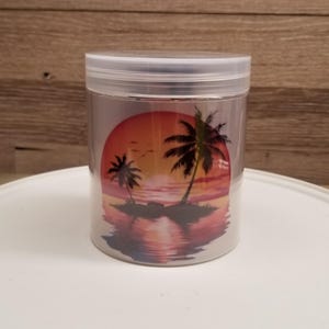 May include: A clear cylindrical plastic container with a screw-top lid. The exterior features a printed graphic of a tropical sunset over the ocean, displaying silhouettes of palm trees on a small island with orange, pink, and purple hues.