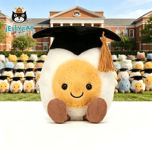 May include: A plush, egg-shaped toy with a smiling face, wearing a black graduation cap with a gold tassel. The toy has a white exterior, a yellow yolk, and brown corduroy feet. Other similar toys are in the background.
