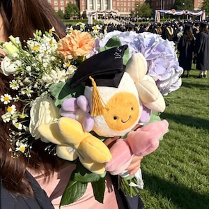May include: A graduation bouquet featuring a plush flower with a mortarboard hat and a smiling face, surrounded by fresh flowers in shades of white, yellow, orange, and purple. The background shows a graduation ceremony.