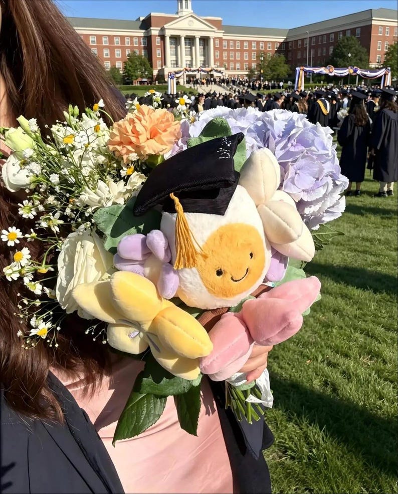 May include: A graduation bouquet featuring a plush flower with a mortarboard hat and a smiling face, surrounded by fresh flowers in shades of white, yellow, orange, and purple. The background shows a graduation ceremony.