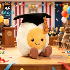 May include: A plush toy egg character wearing a graduation cap with a gold tassel, sitting on a wooden stage. The egg has a smiling face and brown corduroy feet. The background includes balloons, streamers, and a graduation ceremony setting.