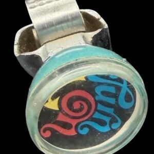 Può includere: Un ditale in metallo color argento con una parte superiore circolare e una copertura in plastica trasparente. All'interno si trova un inserto nero con una spirale rossa e la scritta "fun" in corsivo blu su sfondo giallo.
