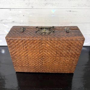 May include: A rectangular, woven basket with a hinged lid. The basket is made of brown wicker and features a diamond pattern on the front. The lid is secured with a metal clasp and hinges.