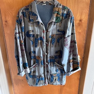 Puede incluir: Una camisa de manga larga con botones en tono gris claro, con un patrón repetitivo de elefantes estilizados en tonos azul, dorado y granate. La prenda incluye un cuello clásico, dos bolsillos delanteros y mangas con puños vueltos.
