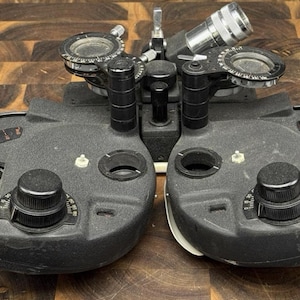 May include: A black ophthalmic phoropter featuring two circular lens housings with adjustable dials, numerical markings, and metallic components. The device includes multiple knobs for precise optical calibration and testing.